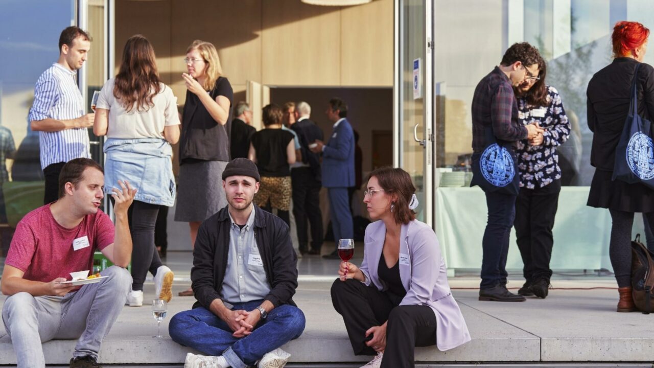 Drei Personen sitzen auf einer Treppenstufe vor einem Universitätsgebäude und unterhalten sich.